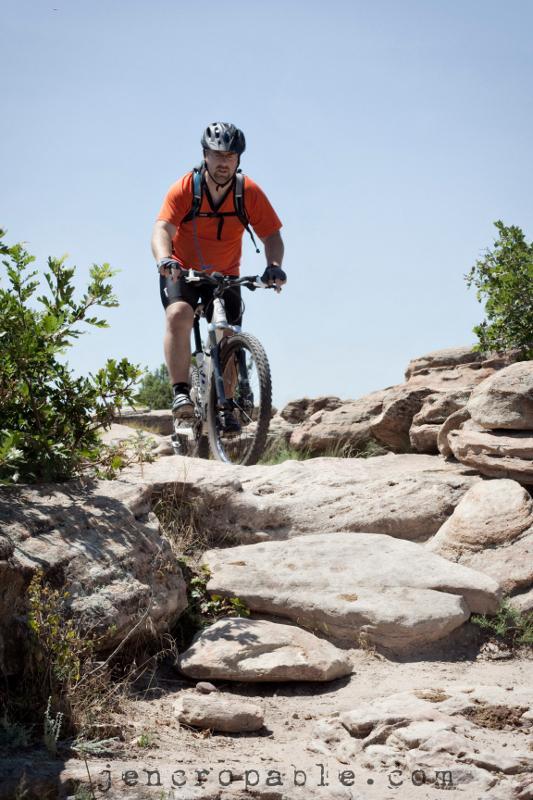 A mountain biker in an orange shirt rides along a rocky trail surrounded by bushes and shrubs under a clear blue sky. Hidden Mesa mountain bike trail.