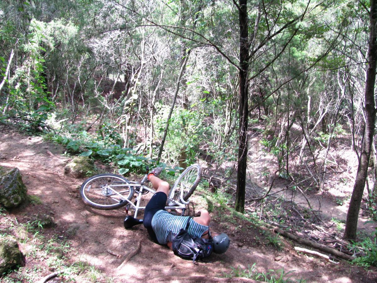 A person lies on their side beside a fallen mountain bike on a dirt trail surrounded by trees and foliage. The individual is wearing a helmet and casual clothing, suggesting they have had an accident while cycling. The scene depicts a wooded area with uneven terrain, highlighting the challenges of off-road biking. Santos mountain bike trail.