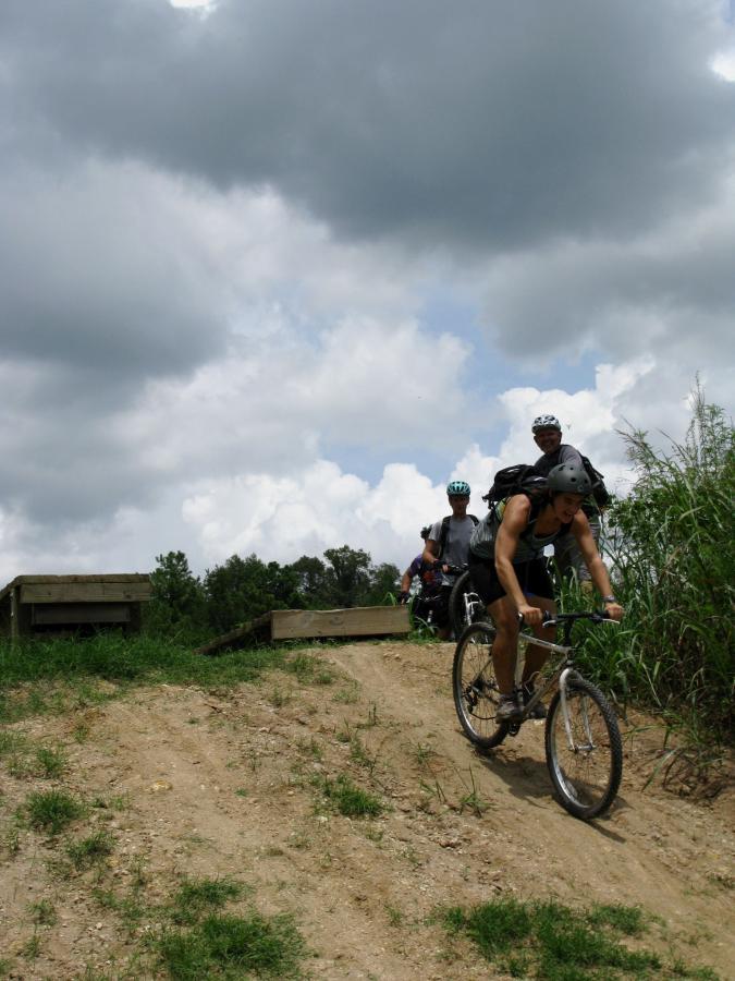 A group of mountain bikers riding on a dirt trail with a ramp in the foreground, under a cloudy sky. One rider, wearing a helmet and biking gear, is descending the ramp, while two others can be seen in the background, preparing to follow. Lush greenery is visible on the sides of the trail. Santos mountain bike trail.