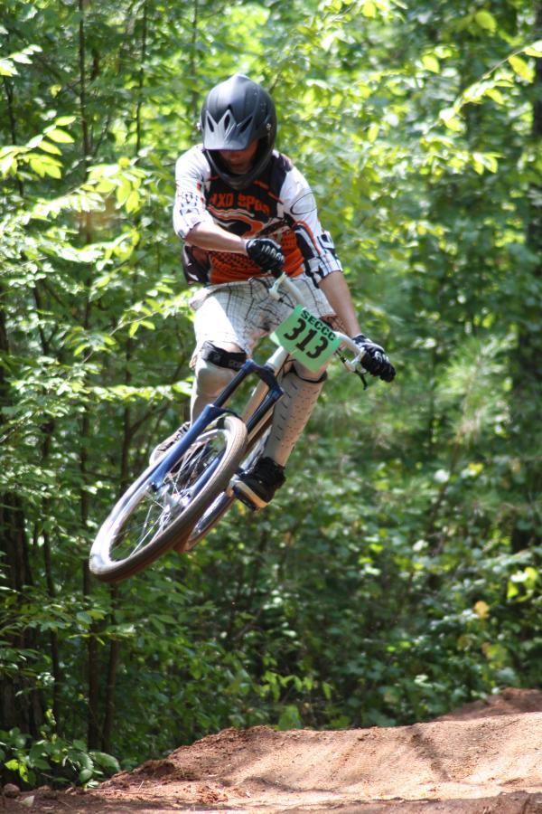 A cyclist in protective gear, including a full-face helmet and padded gloves, performs a jump on a mountain bike over a dirt ramp in a forested area, surrounded by lush green foliage. The bike has a visible race number, and the bike's front wheel is lifted off the ground, emphasizing the action. Georgia International Horse Park mountain bike trail.