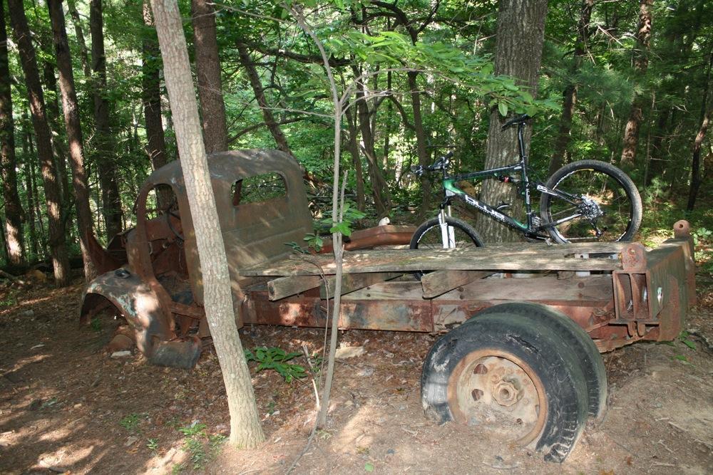 An old, rusted truck partially hidden in a forest, with a mountain bike resting on its flatbed. Surrounding trees and foliage create a lush, natural setting. Bull / Jake Mountain mountain bike trail.