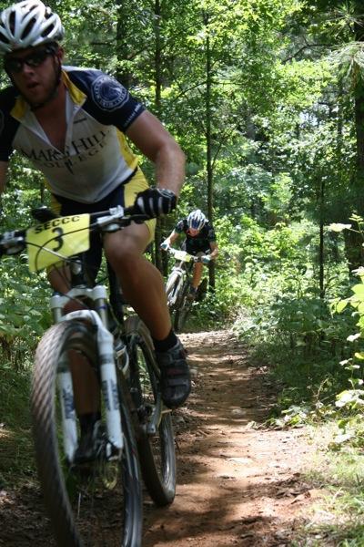 A pair of mountain bikers racing along a dirt trail surrounded by lush greenery. The lead rider is in the foreground, wearing a yellow and blue jersey, while a second rider trails closely behind. Sunlight filters through the trees, highlighting the vibrant environment. Georgia International Horse Park mountain bike trail.