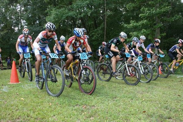 A group of competitive mountain bikers lined up at the starting line in a grassy area, preparing to race. They are wearing colorful cycling jerseys and helmets, some with race numbers visible on their bikes. A few orange cones are placed near the starting point, and trees can be seen in the background. Georgia International Horse Park mountain bike trail.