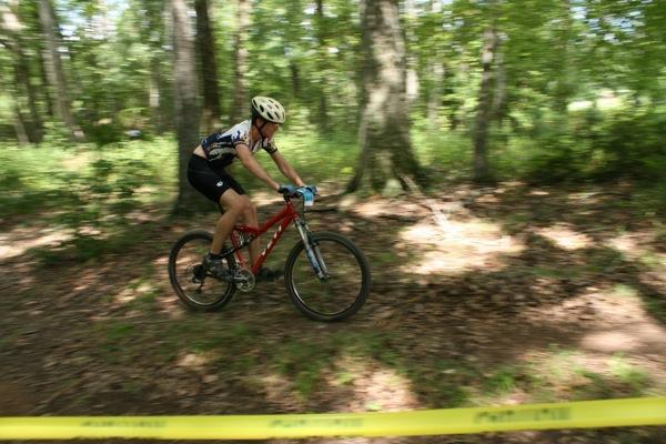 A cyclist in a helmet rides a red mountain bike along a dirt trail surrounded by trees. Sunlight filters through the leaves, casting dappled shadows on the ground. Yellow caution tape is visible in the foreground, indicating a boundary along the trail. Georgia International Horse Park mountain bike trail.