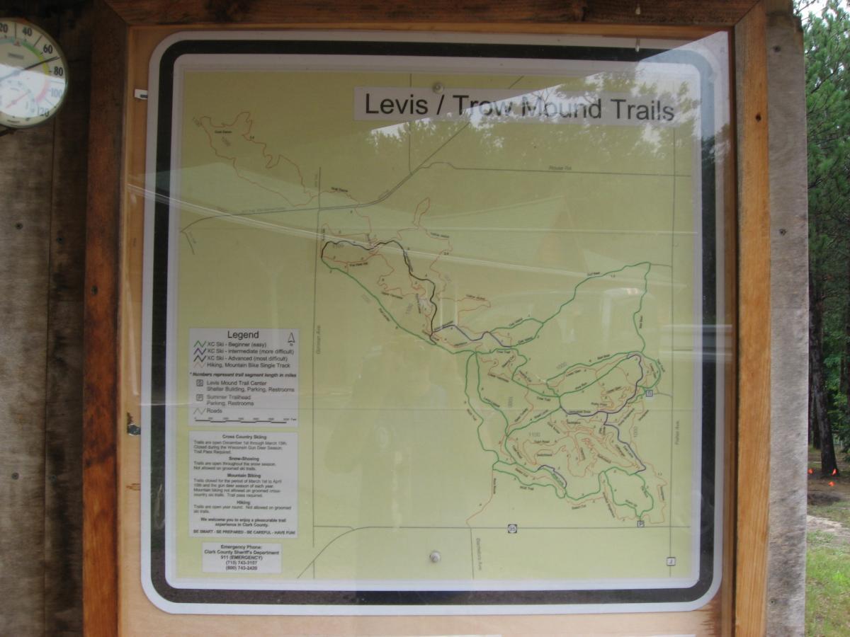 Map of the Levis/Trow Mound Trails, showing various trail routes and difficulty levels, surrounded by a wooden frame. Includes a legend for trail classifications, parking information, and emergency contact details. A thermometer is visible in the corner. Levis Mounds mountain bike trail.