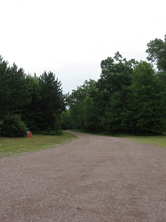 A gravel path winding through a lush green forest, with trees on either side and a cloudy sky overhead. A red trash can is visible on the left side near the edge of the path. Levis Mounds mountain bike trail.