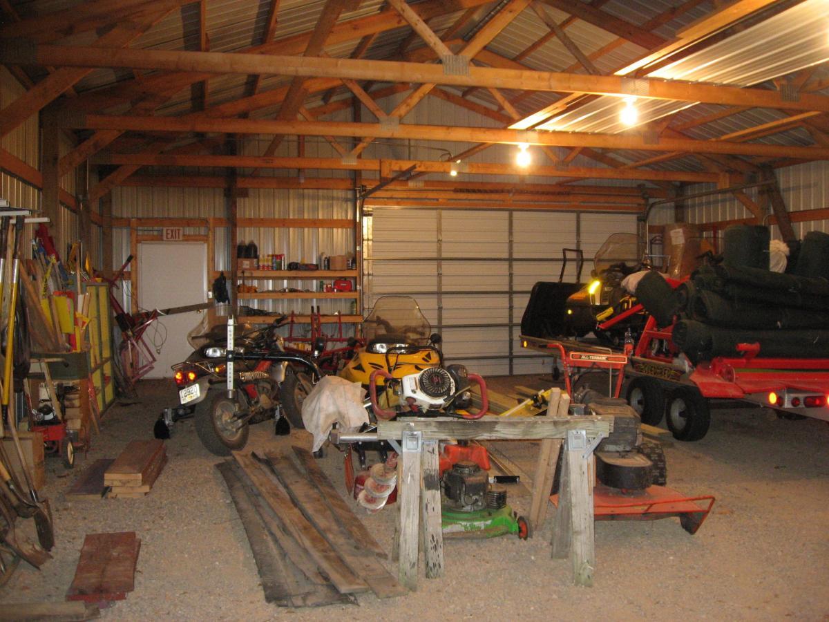 A cluttered garage interior featuring wooden beams and various tools. On the left, shelves hold equipment and a bicycle, while garden tools lean against the wall. In the center, a motorcycle and a yellow all-terrain vehicle are parked beside a workbench. On the right, a red trailer is loaded with equipment, including green rolls, indicating a space for both vehicle storage and home maintenance activities. Soft lighting illuminates the area, creating a functional yet busy atmosphere. Levis Mounds mountain bike trail.