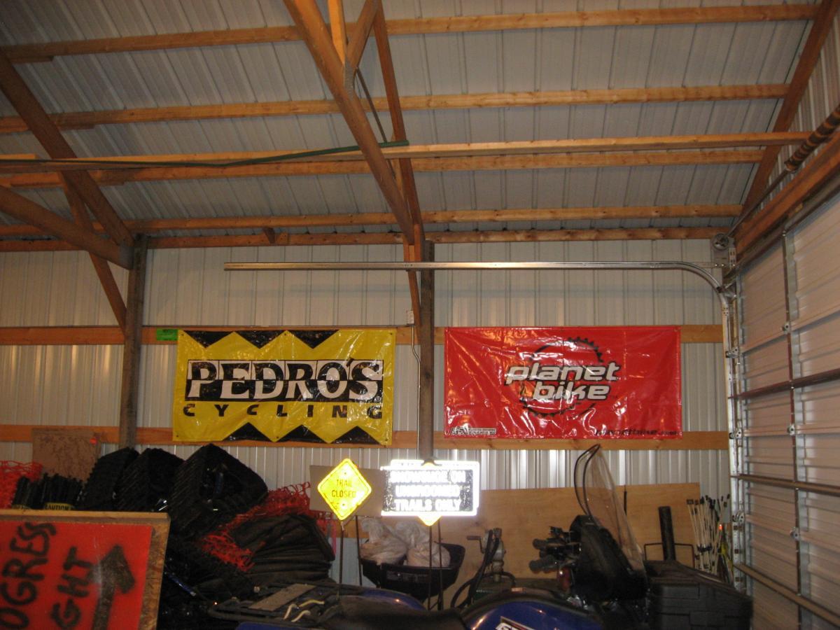 Interior of a garage displaying two large banners: one yellow and black banner for "Pedro's Cycling" and another red banner for "Planet Bike." The scene also includes stacked items and a partially visible sign. The ceiling features wooden beams, contributing to the rustic atmosphere of the space. Levis Mounds mountain bike trail.