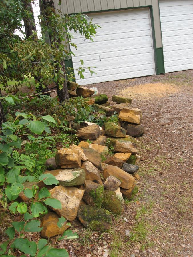 A rocky stone wall partially covered with moss, lined with greenery, beside a gravel path leading to a garage with white doors. Levis Mounds mountain bike trail.