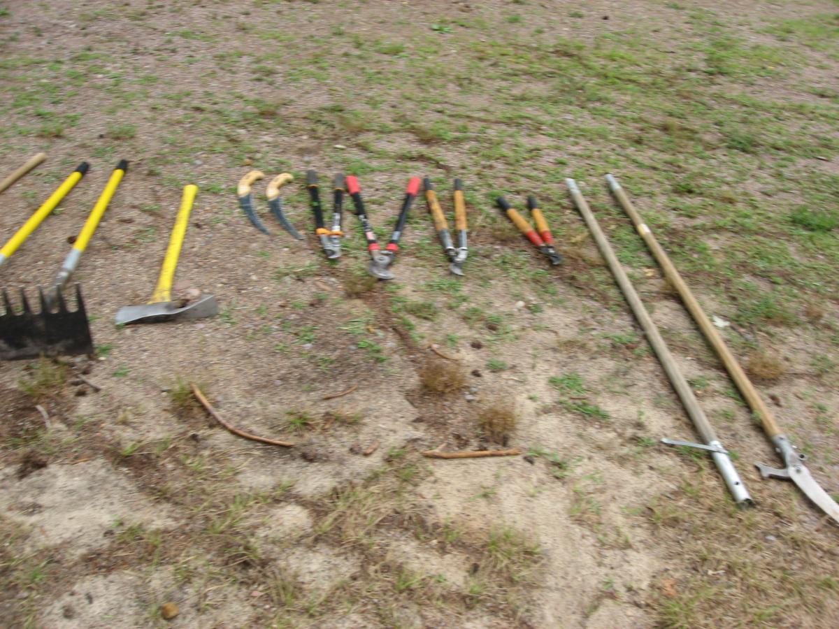 A collection of gardening and landscaping tools arranged on a patch of dirt, including a rake, several shovels, pruning shears, and hoes, set against a background of sparse grass and soil. Levis Mounds mountain bike trail.