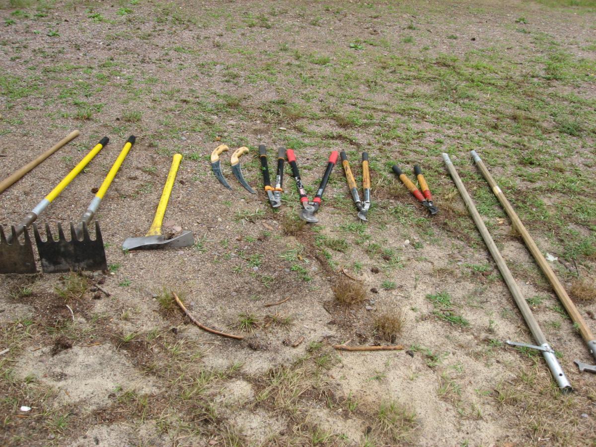 A variety of gardening tools arranged on the ground, including rakes, shovels, pruning shears, and hoes, set against a backdrop of dirt and sparse grass. Levis Mounds mountain bike trail.