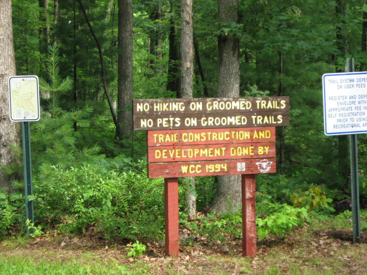 A wooden sign in a forested area stating "No Hiking on Groomed Trails, No Pets on Groomed Trails," with additional text about trail construction and development by WCC in 1994. There is also a blue informational sign nearby. Green foliage and trees surround the signs. Levis Mounds mountain bike trail.