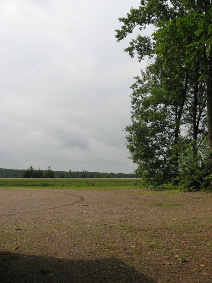 A tranquil outdoor scene featuring a gravel area bordered by lush green trees on the right and a grassy field extending into the distance under a cloudy sky. Levis Mounds mountain bike trail.