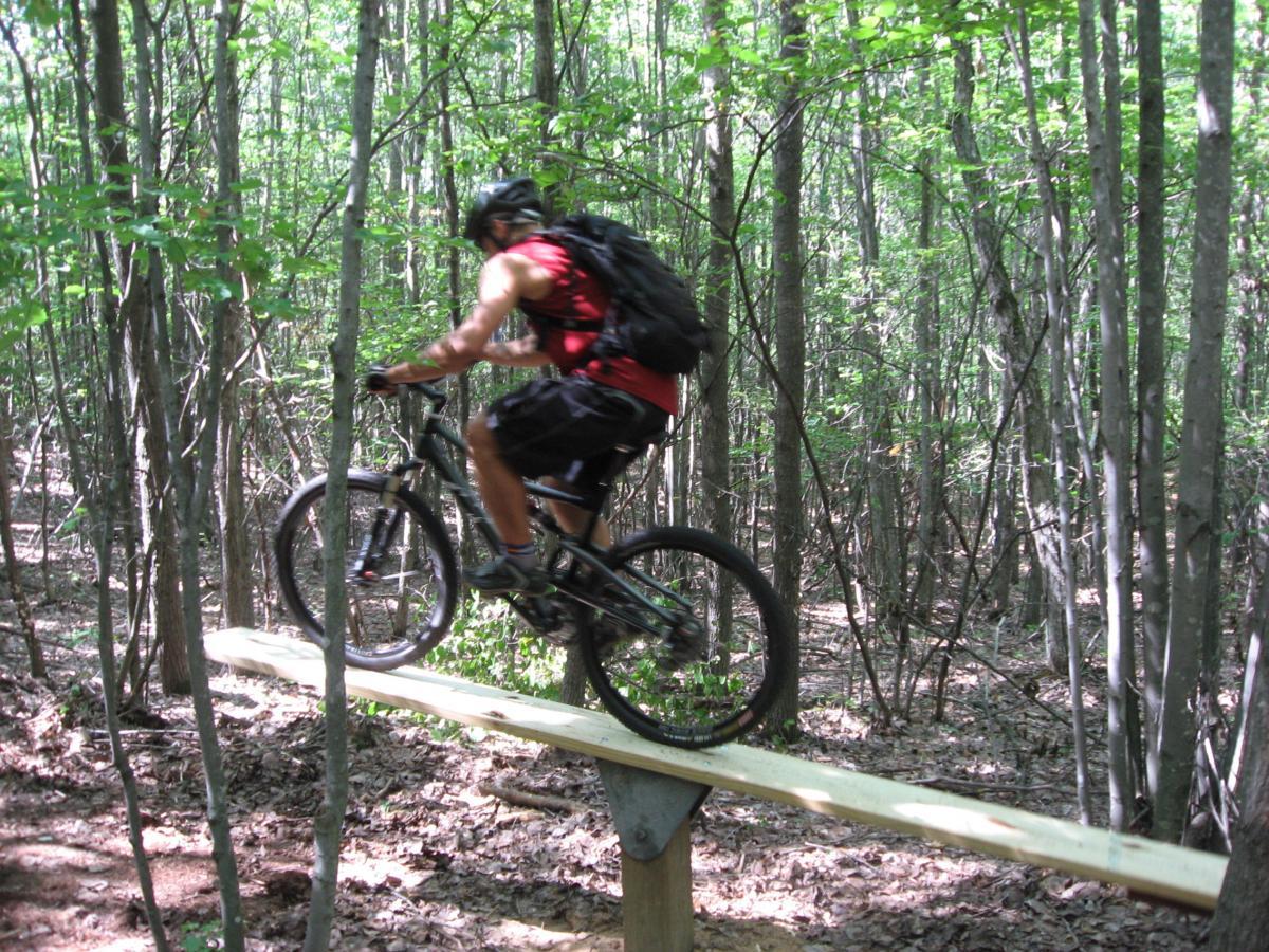 A mountain biker navigating a wooden plank bridge in a dense forest, surrounded by trees and foliage. The cyclist is wearing a helmet and a backpack, showcasing action and skill as they balance on the narrow path. Levis Mounds mountain bike trail.