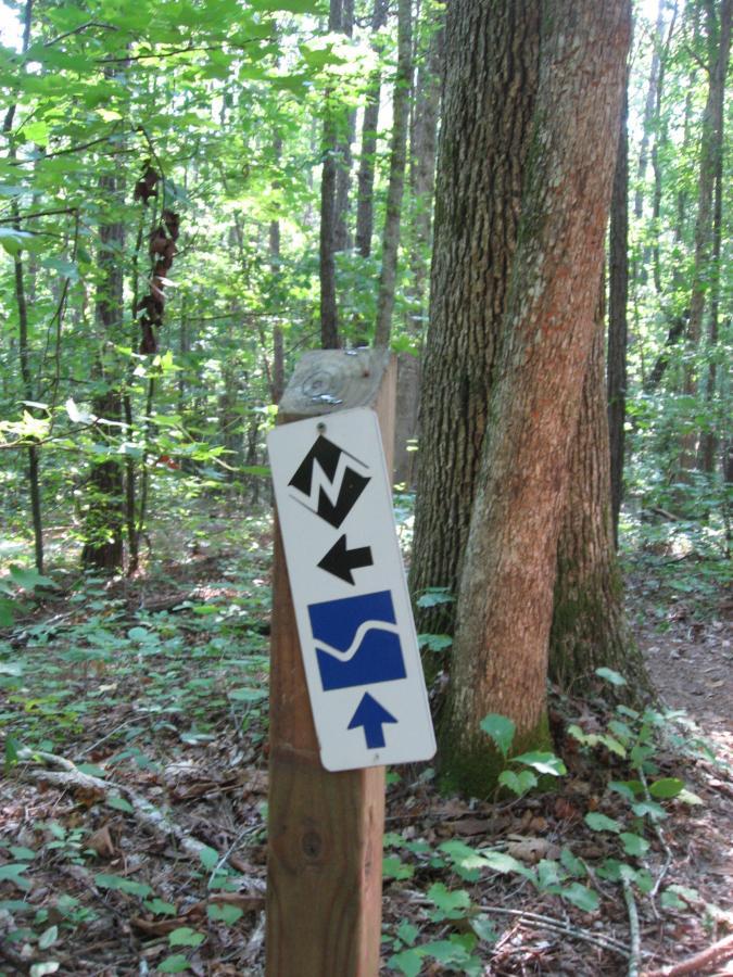 A trail sign in a wooded area, featuring directional arrows. The sign indicates North, with an arrow pointing up, a left arrow, and a wavy line symbol below. Surrounded by green foliage and tall trees, the sign is mounted on a wooden post amidst a natural setting. Heritage Park mountain bike trail.