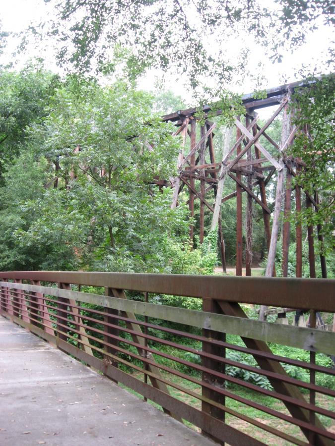 A view of a weathered wooden railroad trestle bridge standing among lush green trees, as seen from a nearby walking path with a railing in the foreground. Oconee River Greenway mountain bike trail.