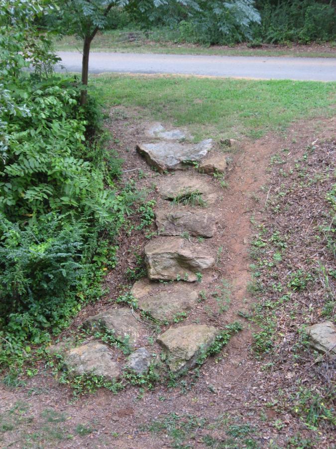 A pathway made of large, natural stones leads through lush greenery, with a dirt trail visible to the right. A tree partially shades the area, and a road can be seen in the background. Oconee River Greenway mountain bike trail.