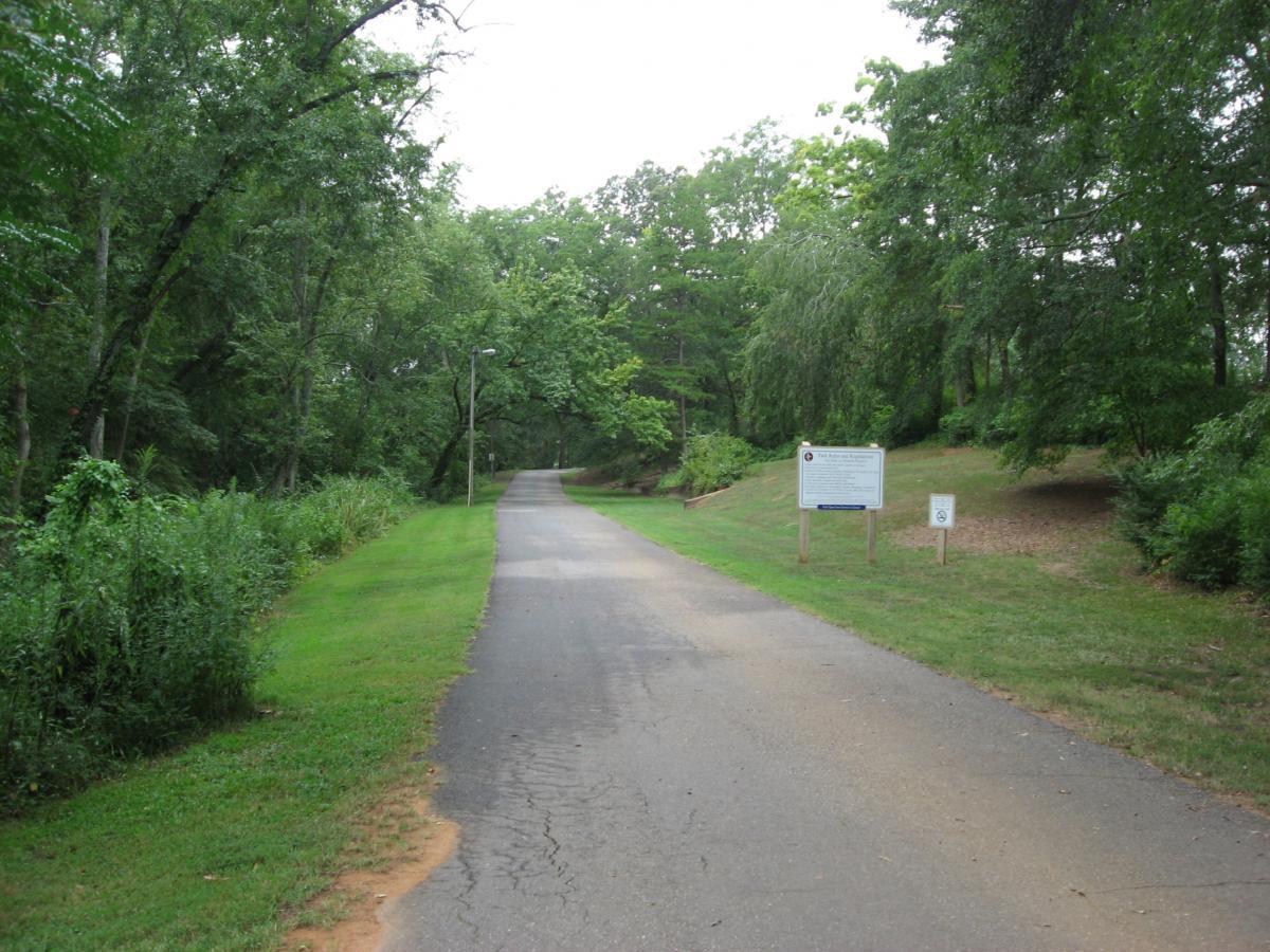 A tranquil pathway lined with lush green trees and foliage, leading into a wooded area. On the right side, there are informational signs beside the road, while the path is paved and appears well-maintained, inviting visitors to explore the natural surroundings. The scene is brightened by gentle overcast skies. Oconee River Greenway mountain bike trail.