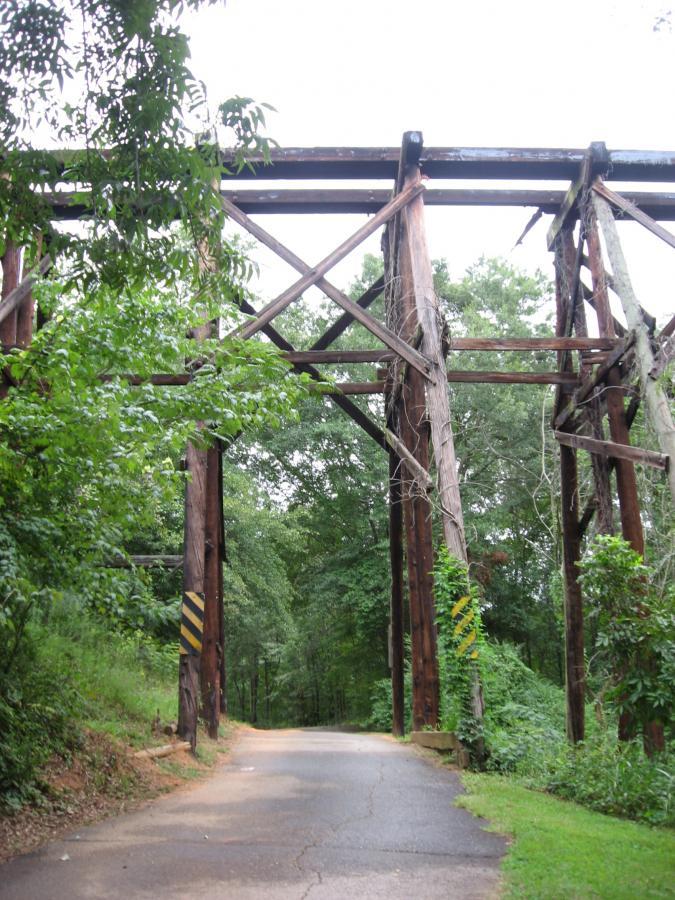 An abandoned wooden bridge structure surrounded by dense greenery, with a narrow paved road leading towards it. The bridge features large support beams and yellow and black striped markings on the sides, indicating caution. The scene is set on a cloudy day, emphasizing the natural surroundings. Oconee River Greenway mountain bike trail.