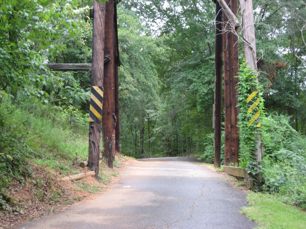 A curved road leading through a wooded area, framed by two large wooden posts with caution signs. Lush greenery surrounds the pathway, creating a natural tunnel effect. Oconee River Greenway mountain bike trail.