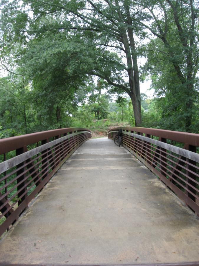 A view of a pedestrian bridge arching over a wooded area, surrounded by lush green trees. The path is made of concrete, and a bicycle is leaning against the railing on the right side. Oconee River Greenway mountain bike trail.