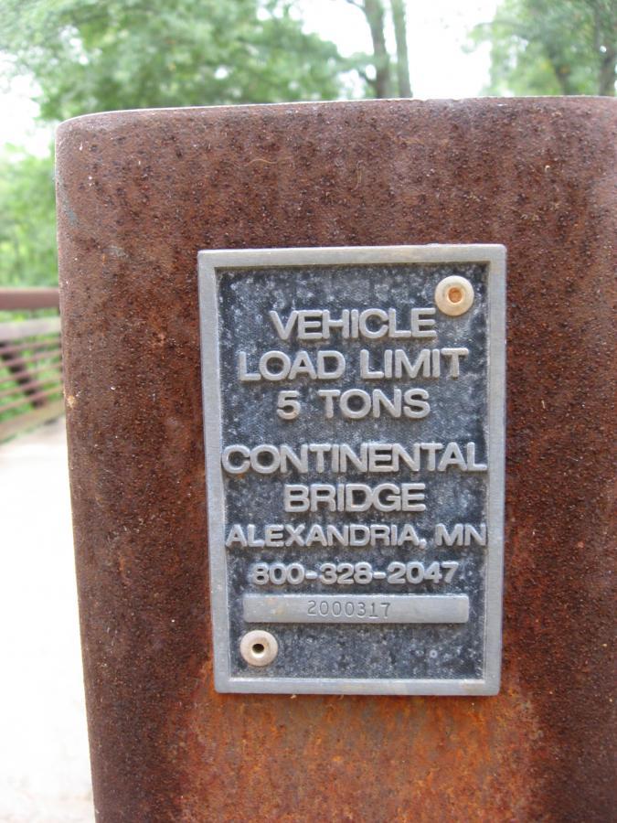 Metal plaque attached to a rusted post displaying the vehicle load limit of 5 tons for the Continental Bridge in Alexandria, Minnesota, along with a contact number and a reference number. The background includes blurred greenery, suggesting an outdoor setting. Oconee River Greenway mountain bike trail.