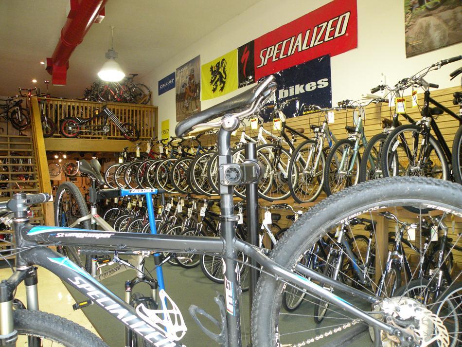 A bike shop interior featuring a variety of bicycles displayed on racks. The foreground shows a black mountain bike on a repair stand, while the background displays an assortment of bikes hanging and arranged on shelves. banners from bike brands are visible on the walls, and a wooden staircase leads to a second level.