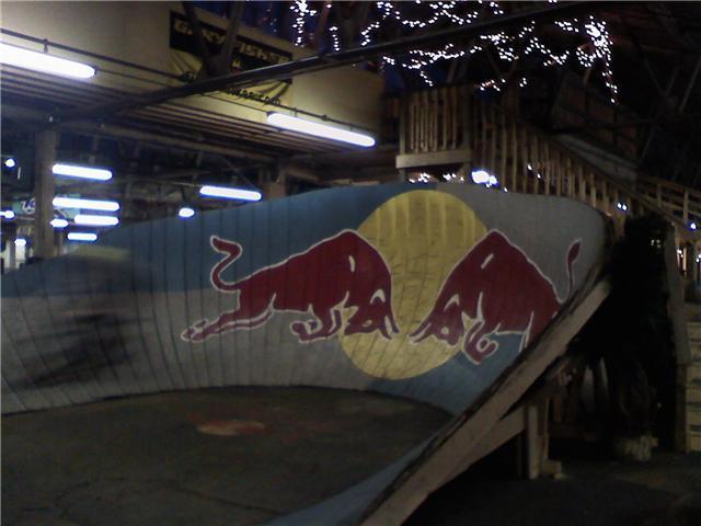 A partially illuminated indoor skate ramp featuring a mural of two red bulls facing each other against a yellow circle, surrounded by wooden structures and decorative lighting. Rays Indoor Mtb Park mountain bike trail.