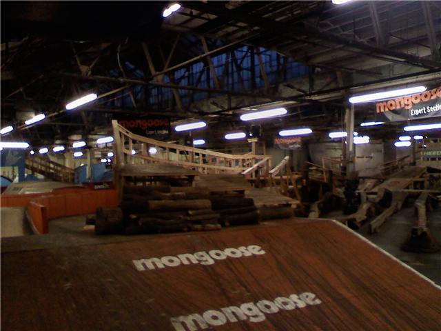 Indoor BMX park featuring various ramps and obstacles. The space includes wooden structures for riders and has signs indicating sponsorship by Mongoose. Bright overhead lights illuminate the area, showcasing the different levels and features available for biking activities. Rays Indoor Mtb Park mountain bike trail.