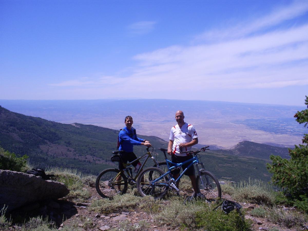 Two mountain bikers stand next to their bikes on a hillside, overlooking a vast valley with greenery and distant mountains under a bright blue sky. The landscape features rolling hills and an expansive view of the terrain below. West Bench Trail mountain bike trail.