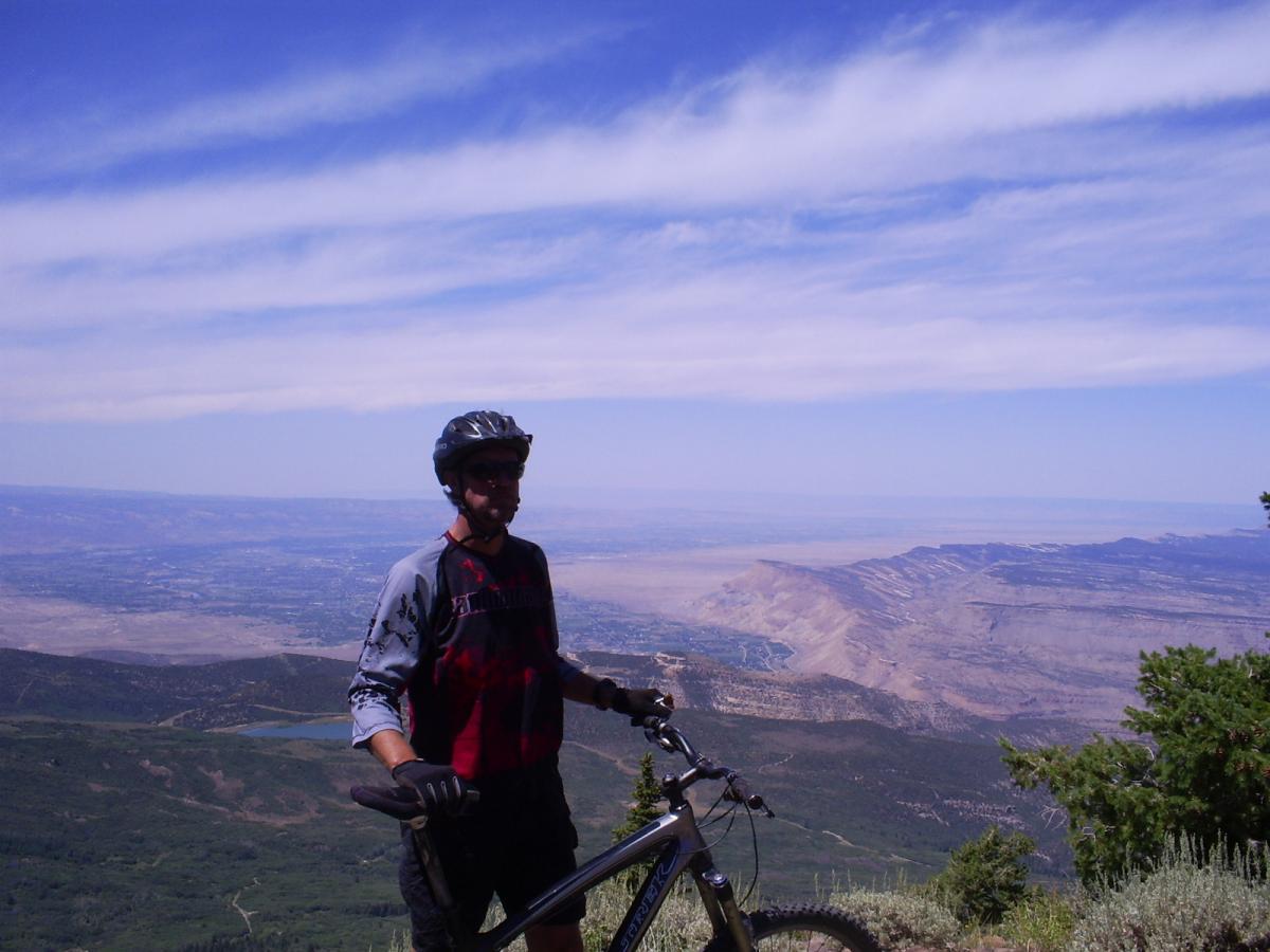 A mountain biker wearing a helmet and gloves stands next to his bike, overlooking a vast landscape of mountains and valleys under a blue sky with wispy clouds. The view includes a distant body of water and rolling hills, showcasing the natural beauty of the area. West Bench Trail mountain bike trail.