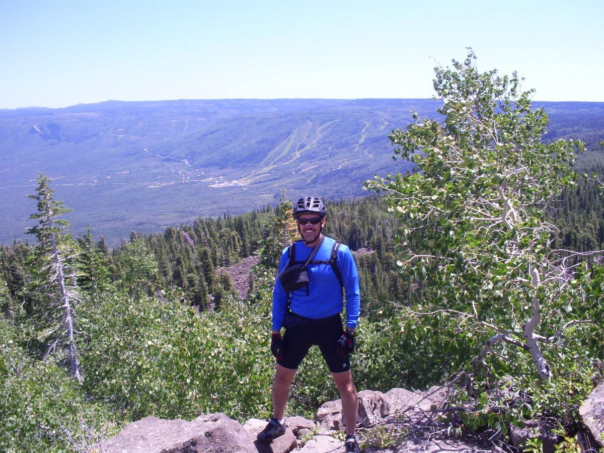 A person wearing a blue long-sleeve shirt, black shorts, and a helmet stands on a rocky outcrop, smiling at the camera. In the background, a panoramic view of a green valley and distant hills is visible under a clear blue sky, with patches of trees in the foreground. West Bench Trail mountain bike trail.