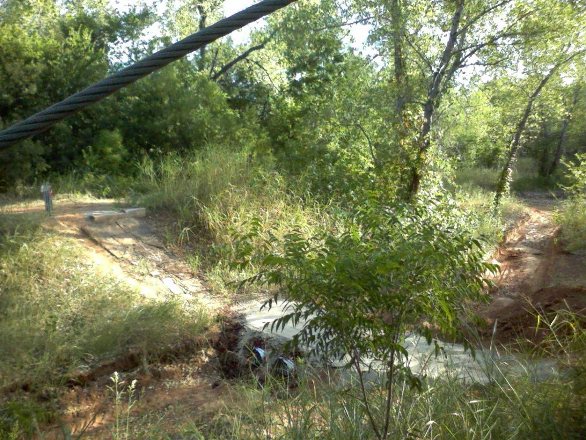 A dirt pathway leads through a lush, green area with tall grass and trees. In the foreground, there's a wide, concrete slab partially visible, suggesting a possible trail or crossing point. The scene is bright and sunny, with foliage providing a natural backdrop. Wee-chi-ta mountain bike trail.