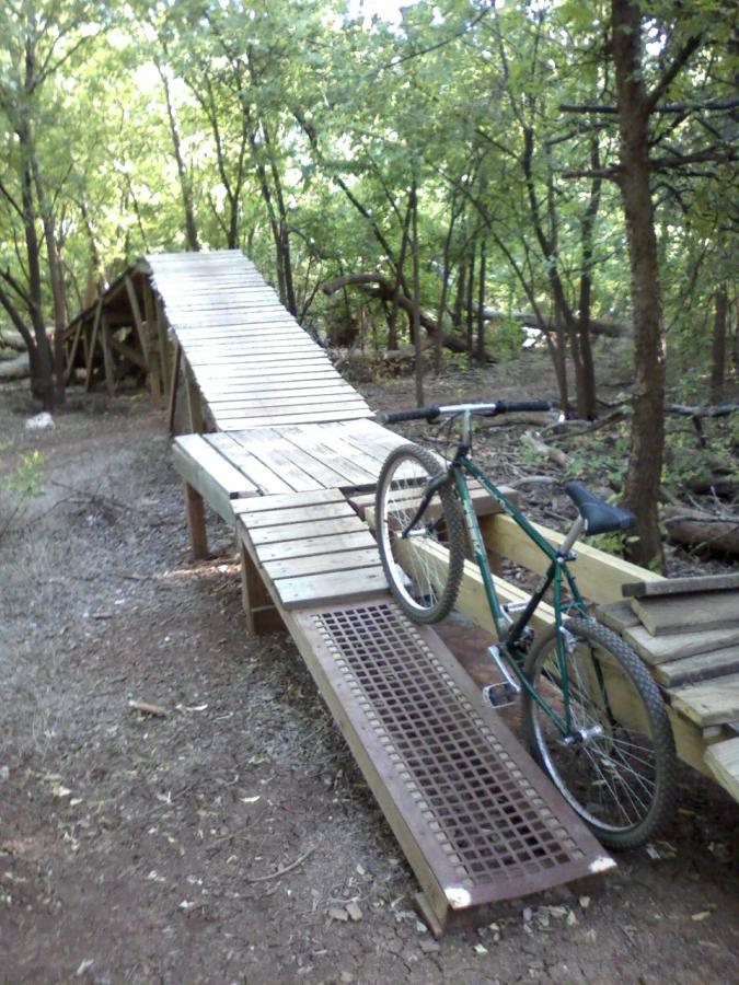 A mountain bike resting on a wooden ramp leading into a forested area, surrounded by trees and natural foliage. The ramp is designed for biking and is made of planks, with a metal grate section visible. The setting indicates a biking trail, with sunlight filtering through the trees. Wee-chi-ta mountain bike trail.