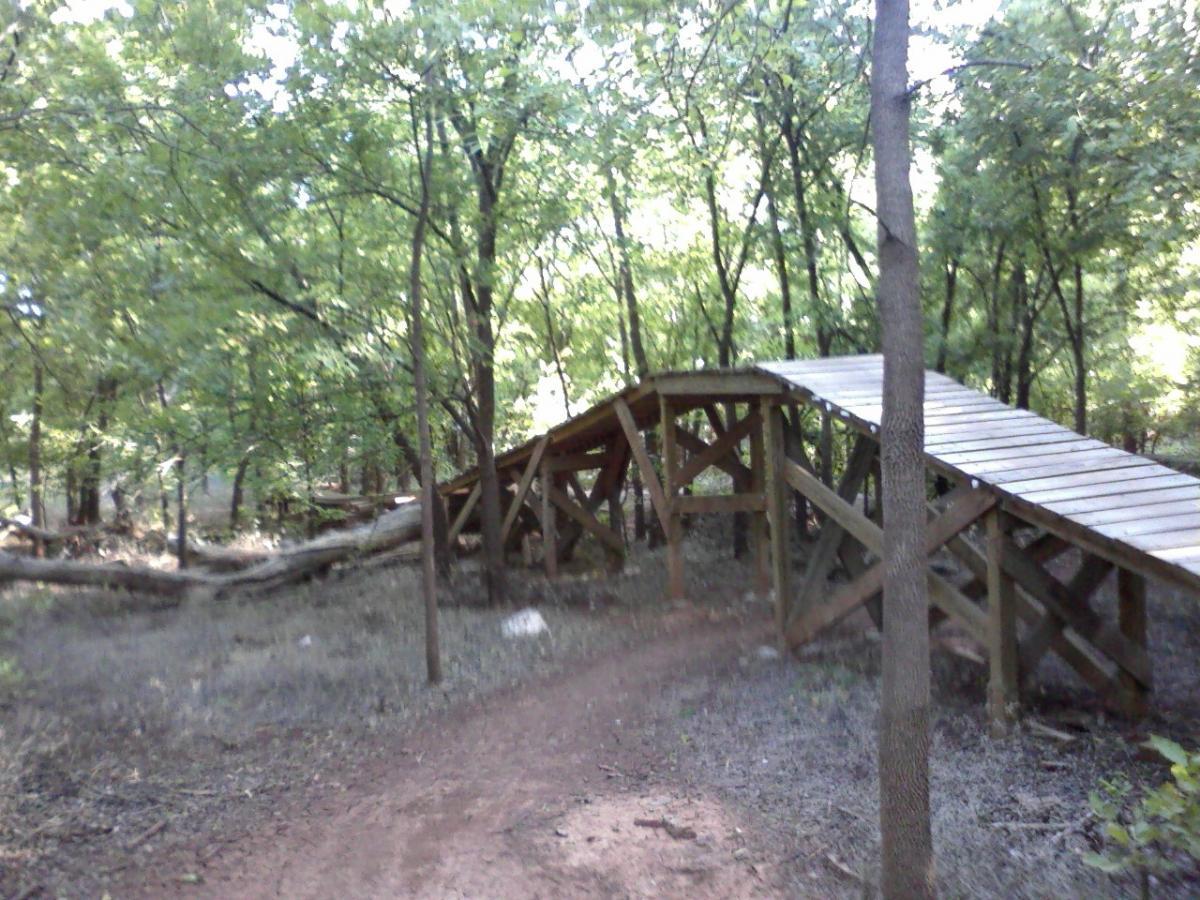 A wooden bridge with a slight incline, set amidst a lush green forest. The pathway is surrounded by trees and thick vegetation, creating a natural, peaceful environment. The sun filters through the leaves, illuminating the scene. Wee-chi-ta mountain bike trail.