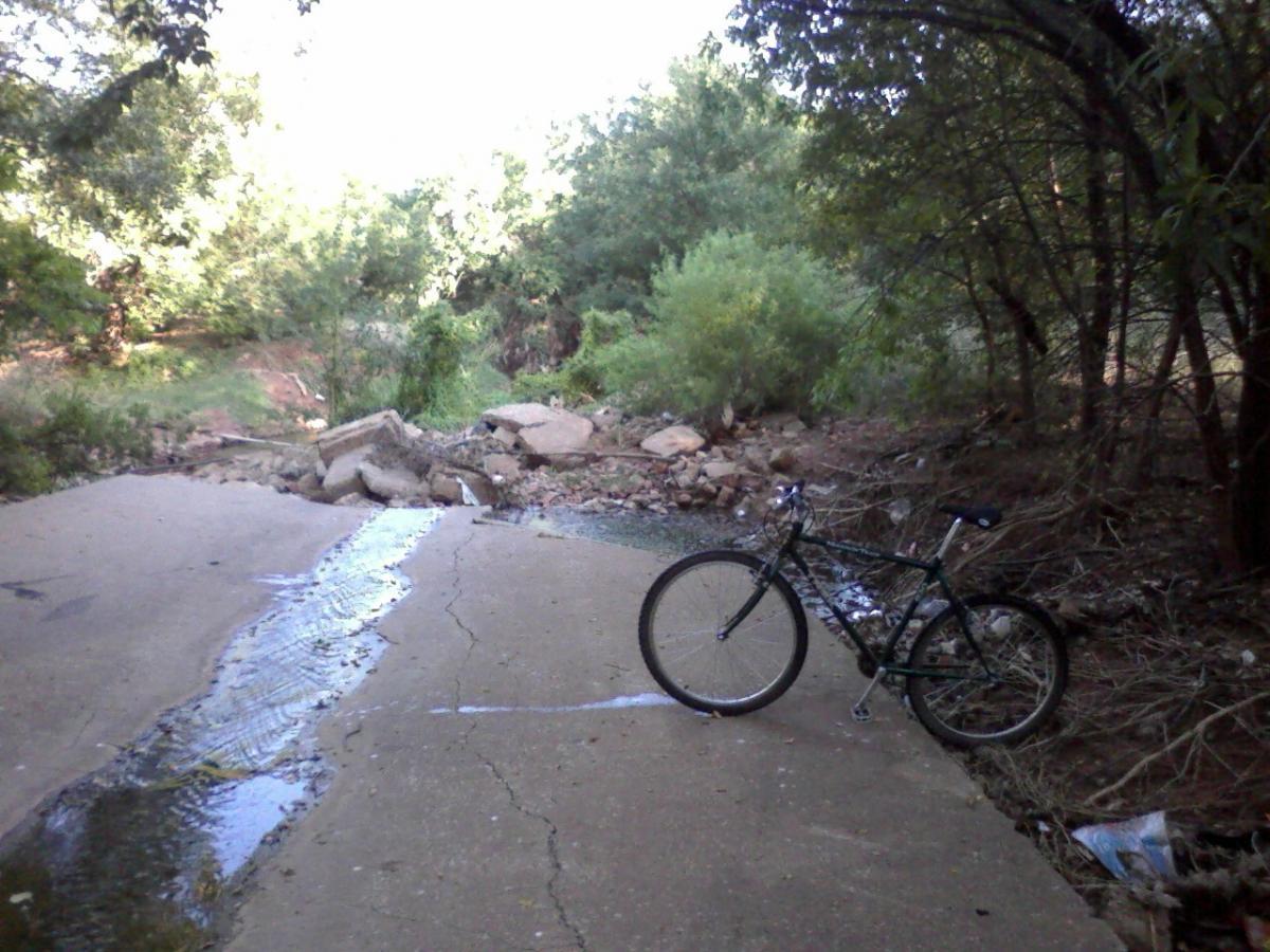 A green bicycle is leaning against a cracked and uneven pavement near a small stream of water flowing through a natural setting. Surrounding the area are trees and shrubs, with some rocks and debris scattered along the path. The scene is peaceful, showcasing the intersection of nature and urban space. Wee-chi-ta mountain bike trail.