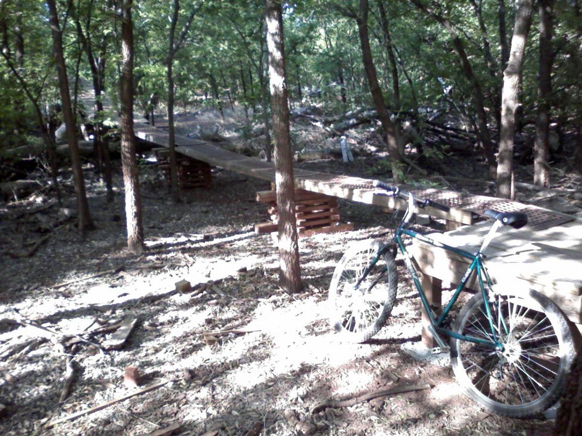 A mountain bike is parked next to a wooden pathway in a wooded area, surrounded by trees and scattered leaves. The sunlight filters through the foliage, illuminating the path and the bike, which has a metallic green frame and visible dirt on the tires, suggesting recent use in the natural terrain. Wee-chi-ta mountain bike trail.
