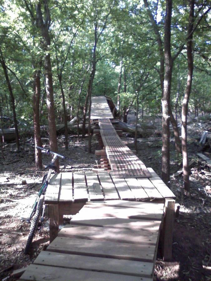 A wooden bike trail winding through a forest, featuring a ramp and bridge made of planks. A mountain bike is parked beside the trail, surrounded by trees and greenery. Wee-chi-ta mountain bike trail.