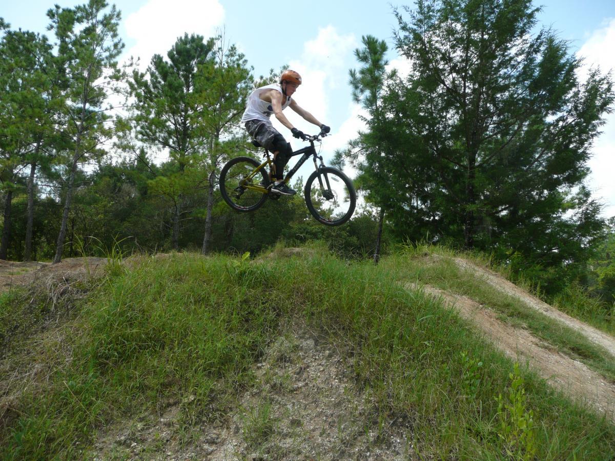 A cyclist wearing an orange helmet performs a jump on a mountain bike over a dirt mound in a wooded area, surrounded by tall green grass and trees under a blue sky. Santos mountain bike trail.