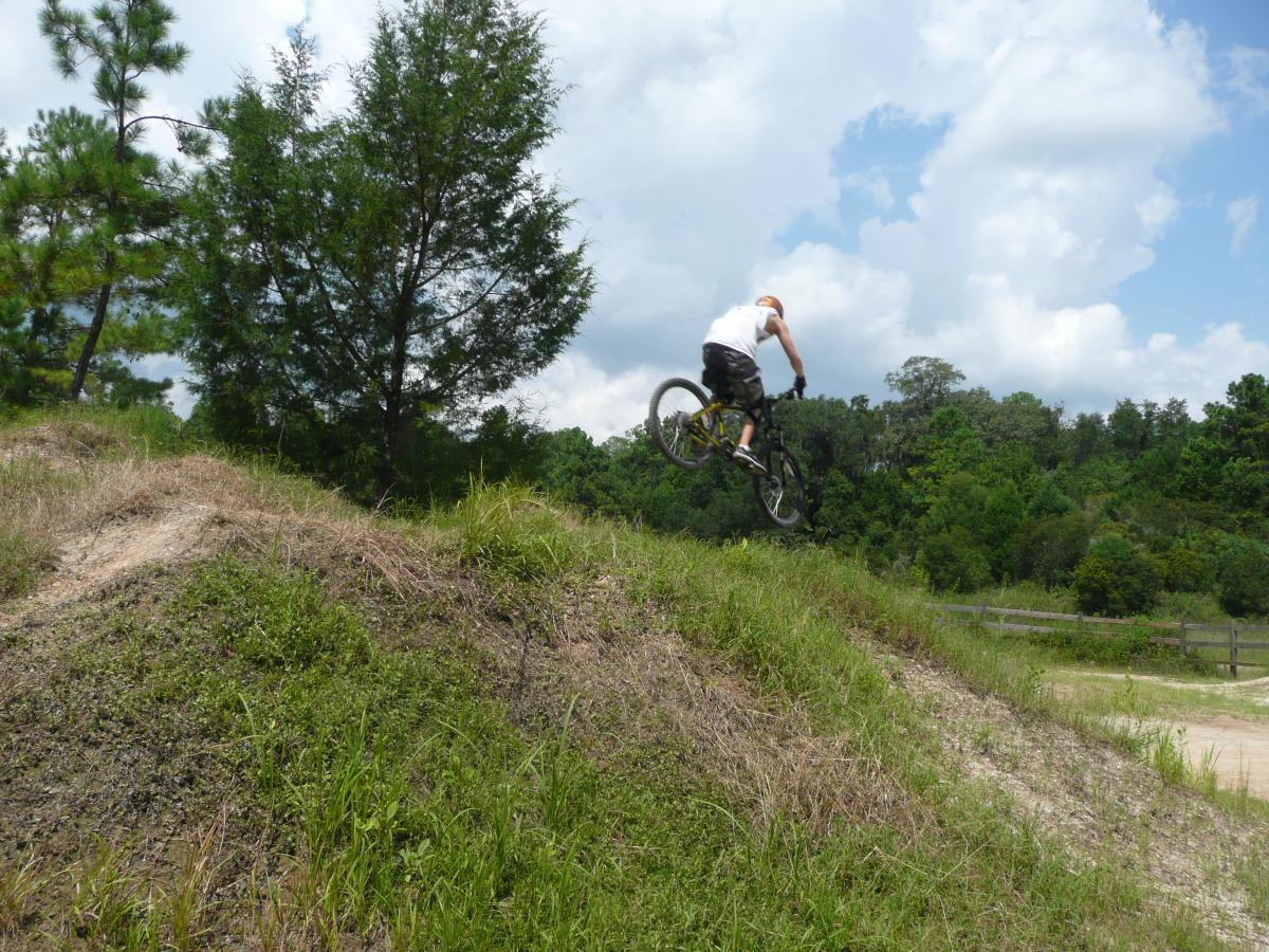 A person wearing a sleeveless shirt and shorts is performing a jump on a mountain bike over a grassy mound, with trees and a blue sky in the background. Santos mountain bike trail.