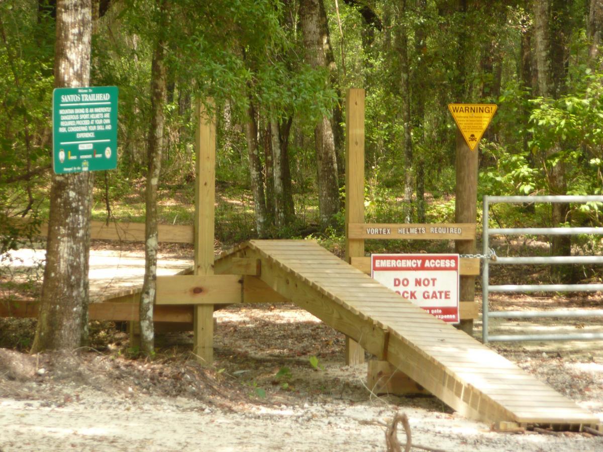 A wooden ramp leads to a trailhead in a wooded area, marked by several signs. The green sign indicates "Santos Trailhead," warning that mountain biking is at one's own risk. A yellow warning triangle highlights the need for Vortex helmets. A red sign states "DO NOT LOCK GATE" for emergency access, with a chain securing the gate in the background. Lush greenery surrounds the entrance, creating a natural atmosphere. Santos mountain bike trail.