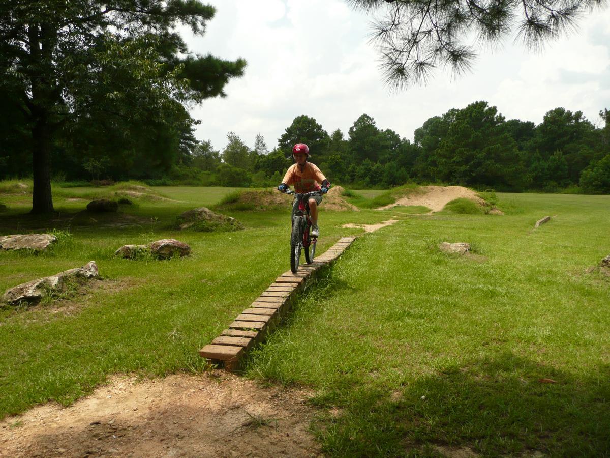 A young person riding a bicycle along a narrow wooden plank bridge in a grassy outdoor area, surrounded by large rocks and trees. In the background, there is a slight hill and some dirt mounds, indicating a biking or recreational park setting. Santos mountain bike trail.