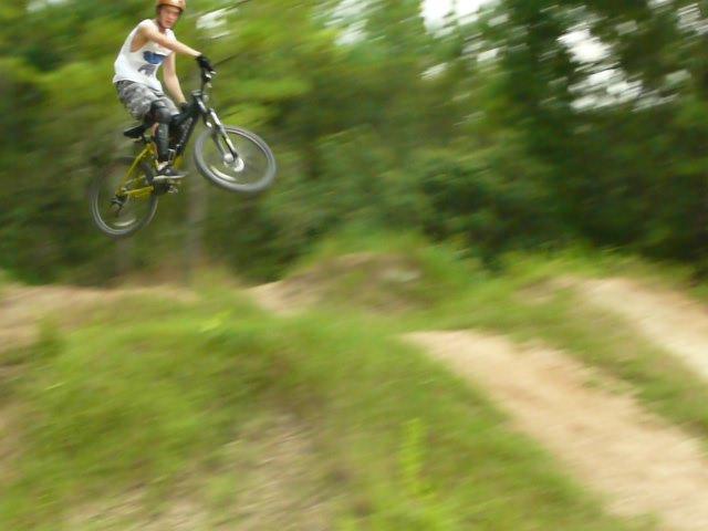 A young man riding a mountain bike performs a jump over a dirt ramp in a green outdoor setting, with trees in the background. The image captures the action, showing the bike in mid-air. Santos mountain bike trail.