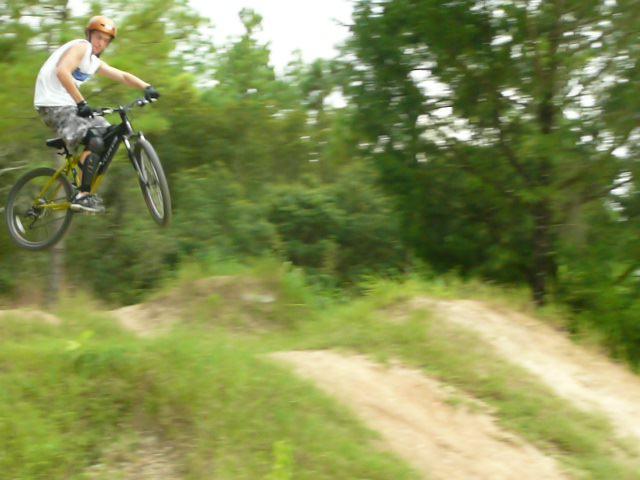 A person wearing an orange helmet and a tank top is performing a jump on a mountain bike over a dirt mound, surrounded by greenery. Santos mountain bike trail.