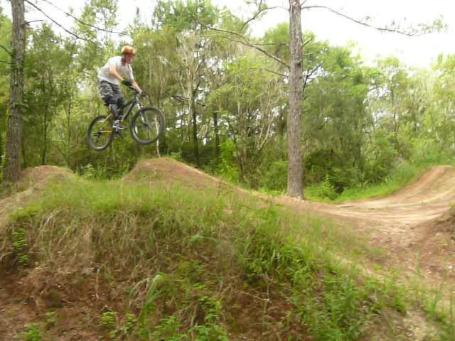 A person in a white shirt and helmet jumps off a dirt ramp while riding a mountain bike, surrounded by trees and greenery in a natural outdoor setting. Santos mountain bike trail.