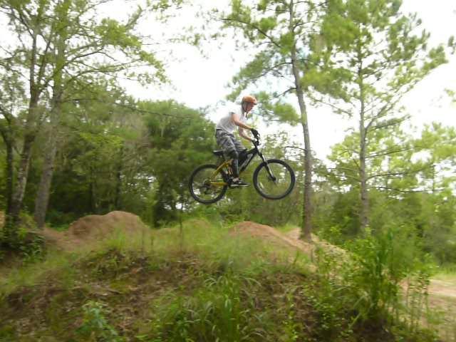 A young rider in a helmet performs a jump on a mountain bike, soaring over a dirt ramp in a wooded area. Tall trees and grassy terrain provide a natural backdrop. Santos mountain bike trail.