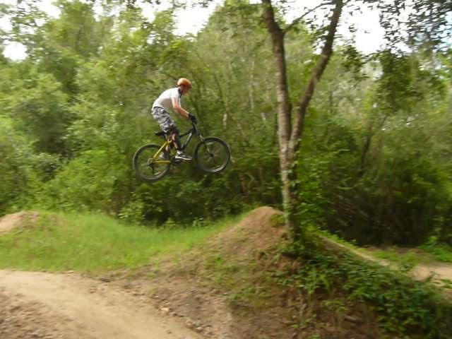 A rider in a helmet jumps off a dirt ramp on a mountain bike, soaring above a grassy area surrounded by trees. Santos mountain bike trail.