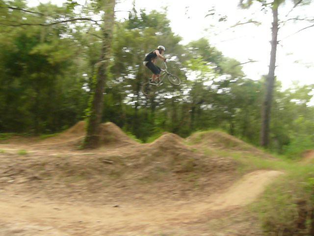 A cyclist performing a jump on a mountain bike over dirt mounds in a wooded area. Santos mountain bike trail.