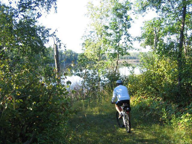 A person riding a mountain bike along a narrow, overgrown path beside a tranquil lake, surrounded by lush greenery and trees on a sunny day. Lake Metigoshe State Park mountain bike trail.