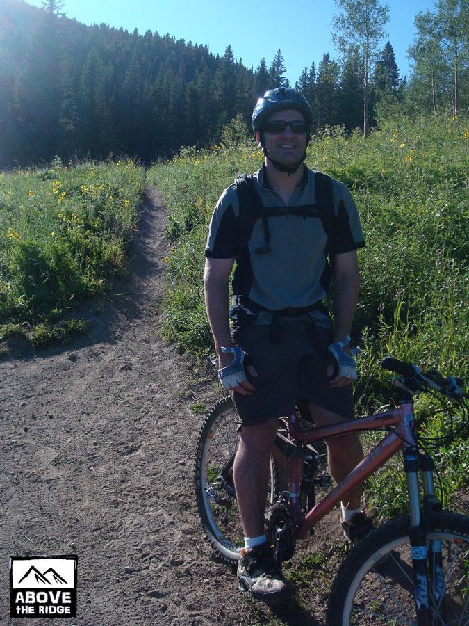 A mountain biker in a helmet and sunglasses stands next to his bike on a scenic trail surrounded by green grass and trees. The sun is shining, indicating a clear day. Snodgrass mountain bike trail.